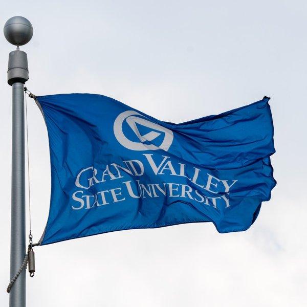 A blue flag flies while on a flagpole. It contains the words Grand Valley State University and the university's logo.