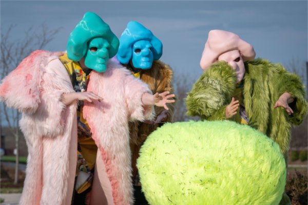 People wearing furry coats of pink, gold and green and wearing masks of green, blue and pink use their hands to gesture while standing in front of a green, furry sphere.