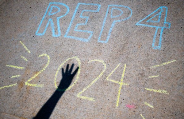 A person's hand showing the number 4 casts a shadow on sidewalk chalk that reads REP4 2024.