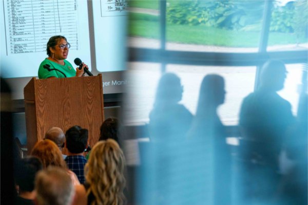  A person speaks at a lectern as people are reflected in glass nearby.