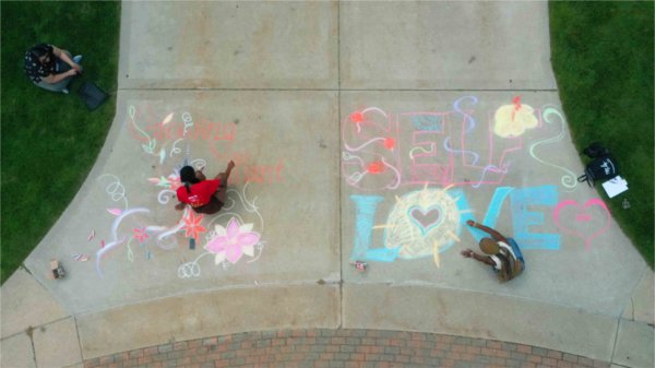  As seen from a drone, students make sidewalk chalk drawings on a sidewalk. 
