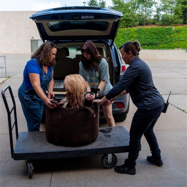 Three people load a plant on a cart into the back of a vehicle.