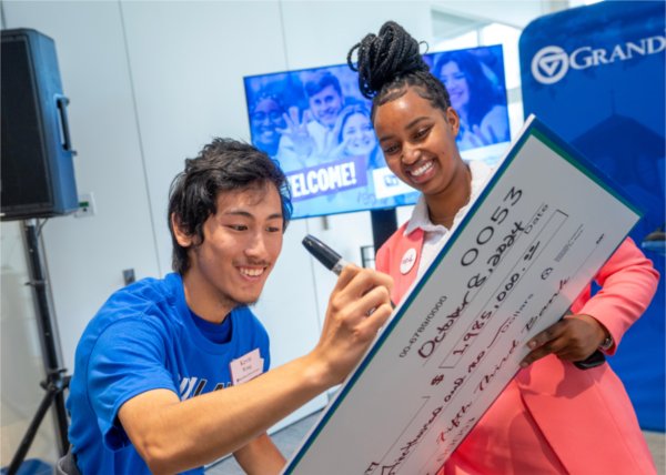 Two GVSU students smile and sign an oversized check made out to Grand Valley State University. 