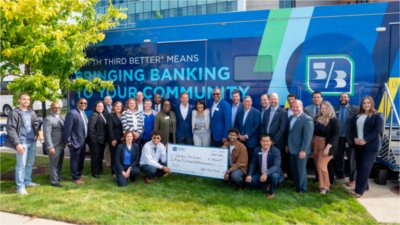 A group of people stand in front of a truck with the Fifth Third Bank logo on it and smile for the camera. Two young men kneel in the middle and hold an oversized check made out to Grand Valley State University.