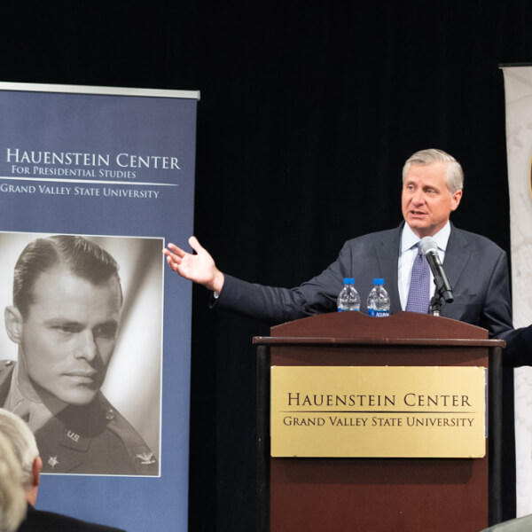 Jon Meacham stands at a podium and gestures to the crowd in the middle of a packed auditorium. The backs of the heads of the audience are blurred in the foreground, Meacham is in sharp focus in the back.