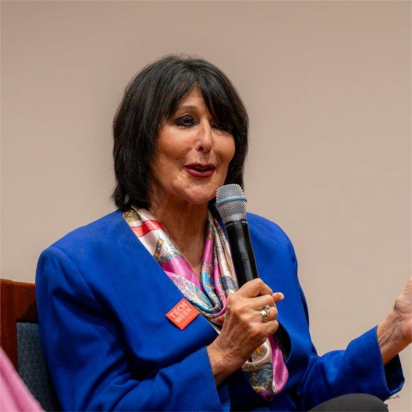 President Mantella speaks into a microphone during a panel discussion. She wears a blue blazer, pink scarf, and a pin that reads �Tech Week�.