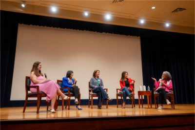 Five women sit in chairs and hold microphones on a small stage. Four of them look to the left at their fellow panel member, who wears a pink blouse and speaks into her microphone.