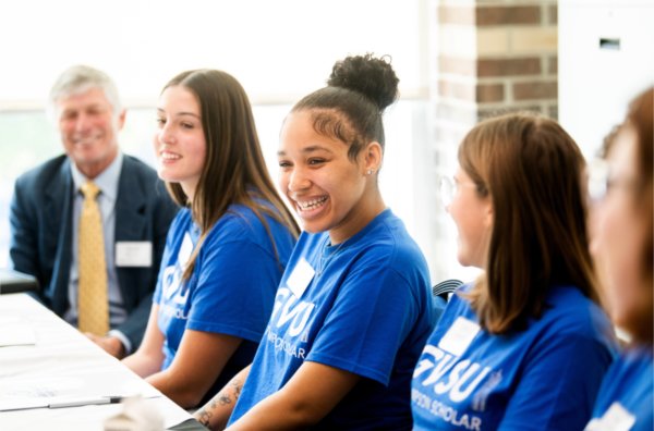 row of students in blue t-shirts, man in suit at end of table