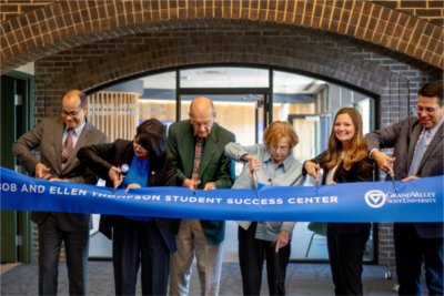 people stand behind a blue ribbon, Bob and Ellen Thompson Student Success Center, with scissors in hand to cut the ribbon