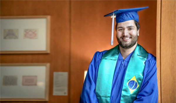 A person wearing academic garb smiles while leaning against a wall.