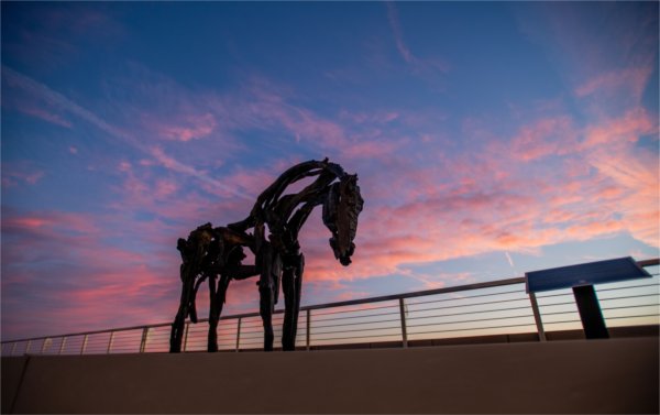 A horse sculpture is next to a railing with a pink sky in the background.