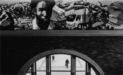 A mural is seen above doors of a building. People are seen walking outside through windows.