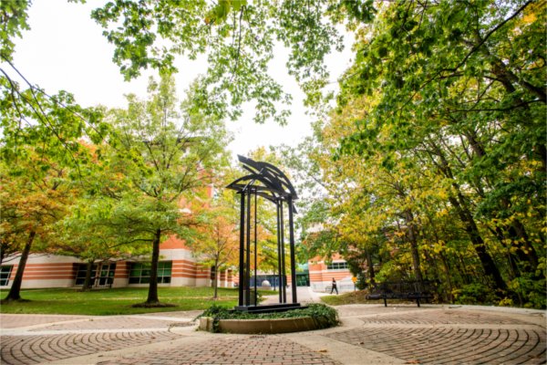 A sculpture sits on a brick walkway, surrounded by trees.