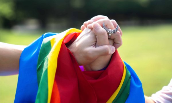  Two hands clasped together are draped in a pride flag.