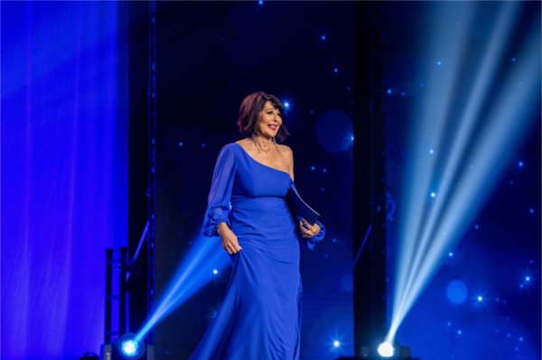  A university president walks onto a stage among blue spotlights wearing a blue evening gown. 