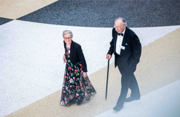  A couple wearing a tuxedo and floral gown are seen from a balcony walking in to an event. 