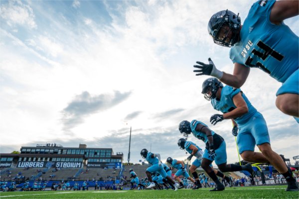 A line of football players run across a football field. They are seen from the ground looking up.