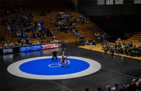 One wrestler on a mat flips the other as a referee and spectators look on.