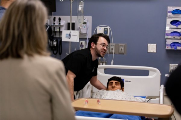 mannequin lying in bed, man in black scrubs leaning over, people looking on