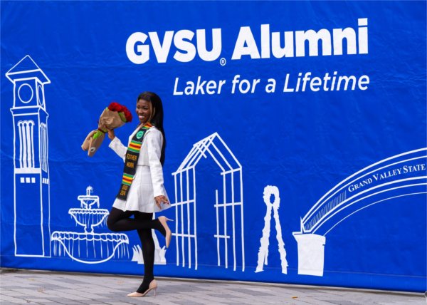 A graduate poses by a "GVSU Alumni Laker for a Lifetime" banner. 