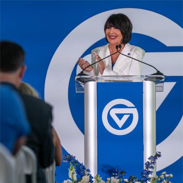 A person at a podium smiles while clapping. The podium and the backdrop both contain the Grand Valley State University logo.