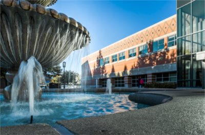 A water fountain sprays water on a sunny day. The Student Services building can be seen behind it.