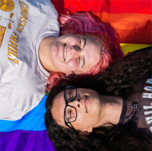 Two people - one with pink hair, the other with long black hair and glasses - lay on a rainbow pride flag and grin up at the camera.