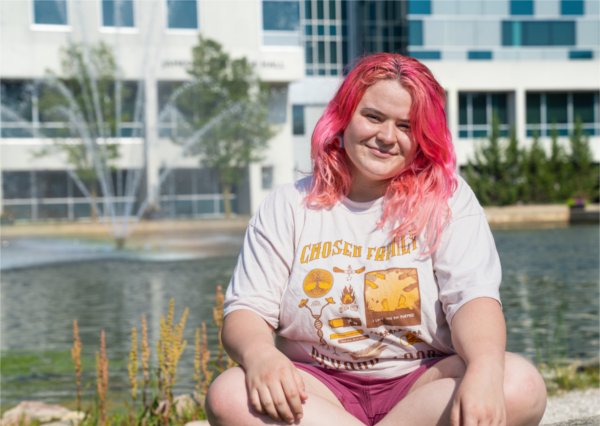 Rowan Armour, a person with pink hair and rainbow earrings, sits in front of Zumberge Pond. They are wearing a shirt that says &ldquo;Chosen Family.&rdquo; 