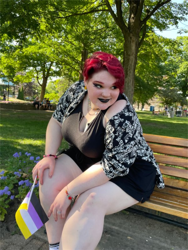 Zoey Belk, a person with short magenta hair, sits on a bench and holds a small non-binary flag with yellow, white, purple, and black stripes.