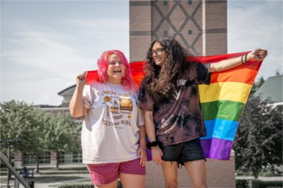 Two people, one with pink hair and the other with long black hair and glasses, hold a pride flag behind them and stand in front of the Cook Carillon clock tower.