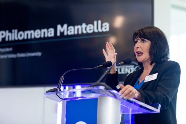 A person holds up a hand while speaking at a podium. The name Philomena Mantella is on a screen in the background.