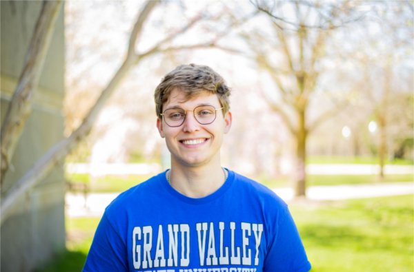 A person smiles for a posed photo with trees in the background. The words "Grand Valley" are visible on the person's shirt.