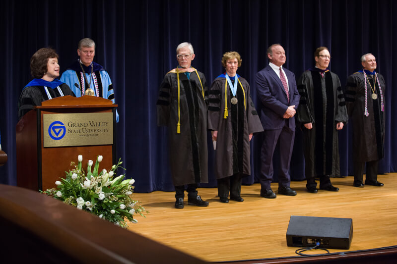 Gayle Davis speaks into microphone; faculty in academic robes to right