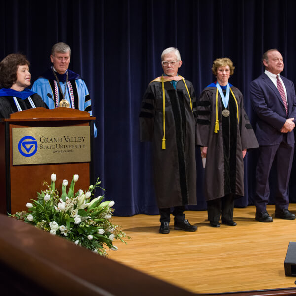 Gayle Davis speaks into microphone; faculty in academic robes to right