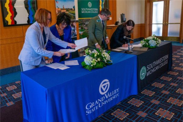 four people signing papers while standing behind two tables with draping for GVSU and SMC