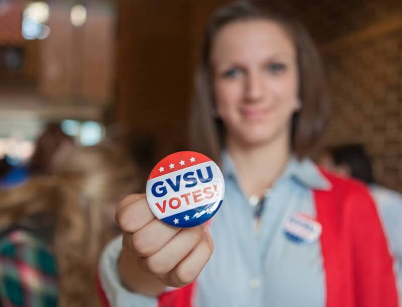 A photo of a student holding up a sticker that says "GVSU Votes!"