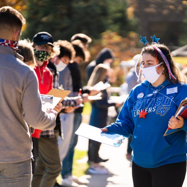 A student in a blue GVSU sweatshirt and holding clipboards hands a clipboard to a student waiting in a line to vote.