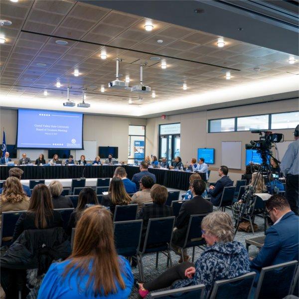 A group of people sit in chairs in a meeting room. Others are sitting at the head table. The screen says, "Grand Valley State University Board of Trustees meeting, February 21, 2025.