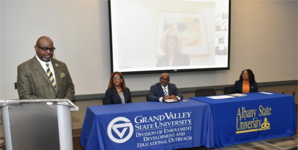 one man at podium with three people seated at table behind with projected screen 