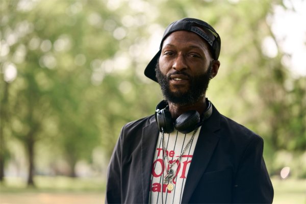 A person wearing a baseball cap backward and with earphones around their neck looks at the camera.