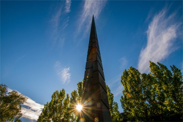  An obelisk sculpture juts into the blue sky as the sun peeks through. 