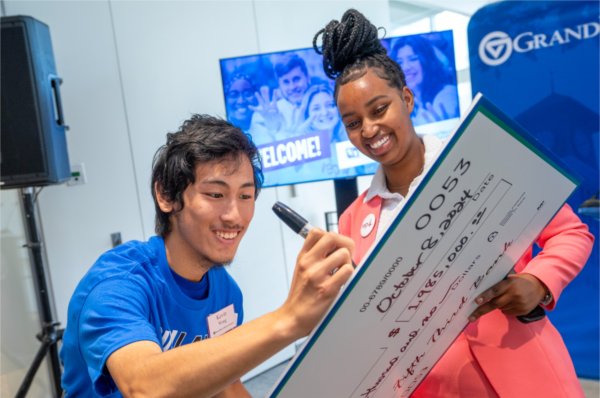  Two smiling students sign an oversized check during a check presentation event. 