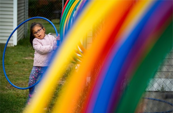  A small child wrestles with a blue Hula Hoop as they try to put it on a rack with a rainbow of colored hoops. 