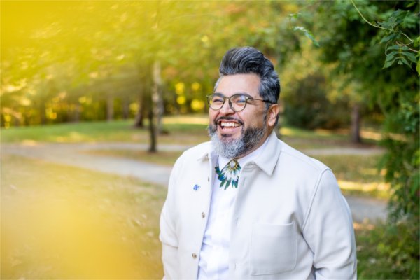   A portrait of a smiling person with glasses wearing a white jacket photographed in a colorful outdoor space. 