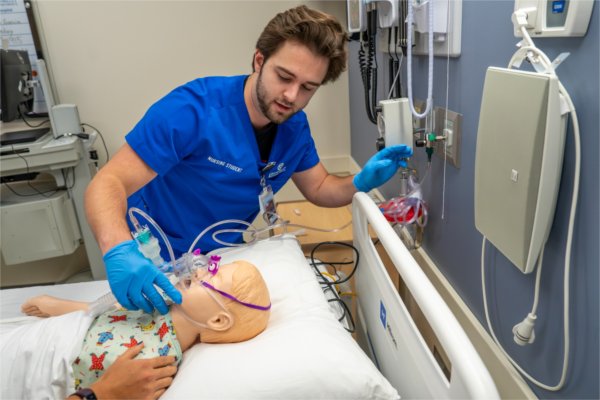  A nursing student wearing a blue scrub top puts an oxygen mask onto a simulation manikin. 