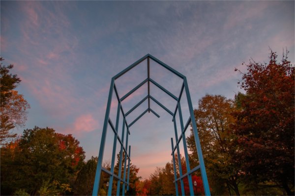  A blue metal sculpture is seen against a pink sunrise and colorful fall leaves. 