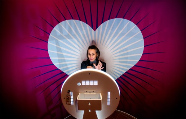  A person gestures while standing in front of a video recording screen with a large pink heart background. 