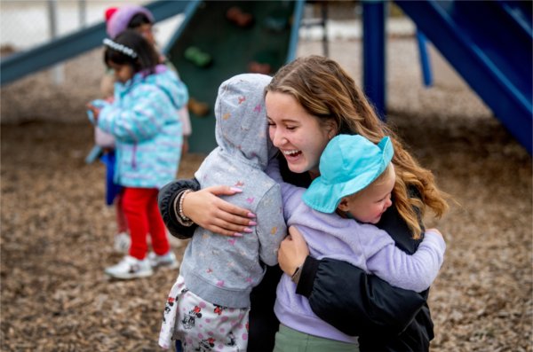  A college student hugs two small children while kneeling on a playground. 