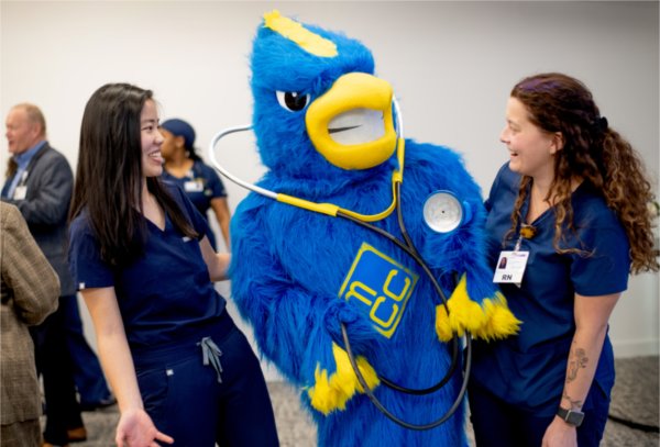 two nursing students in dark blue scrubs laugh with a jaybird mascot in center with a stethoscope around its neck