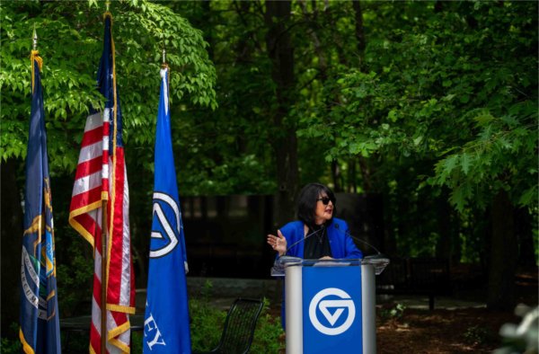 A person standing at a podium with a GVSU logo gestures with one hand while looking off to the side and speaking. Three flags are to the person's right.